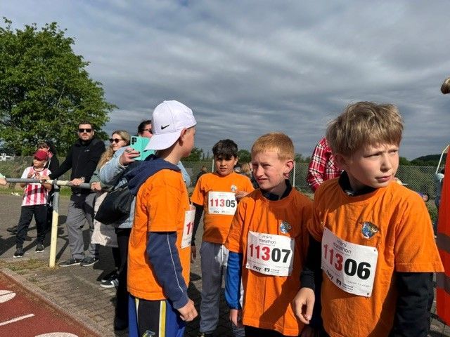 Runners in orange shirts gather at a race start line outdoors under cloudy skies.