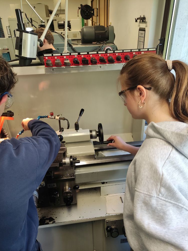 Two people operating a metal lathe in a workshop, with tools and machinery in the background.