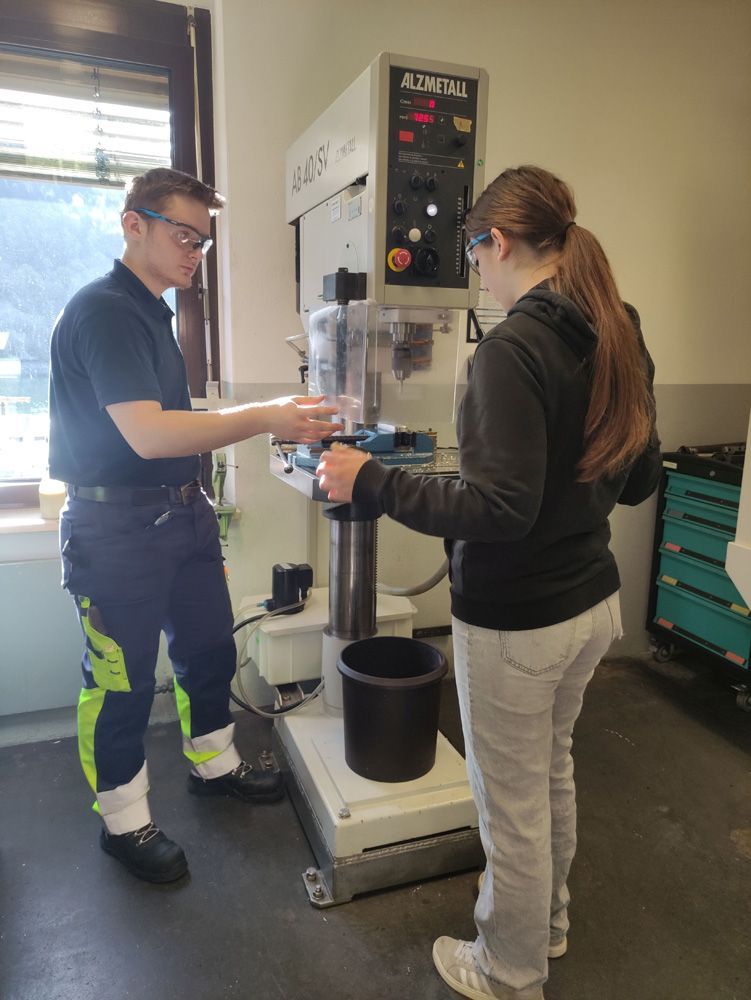 Two people operate a lab machine in an industrial workspace, with tools and a black bucket nearby.
