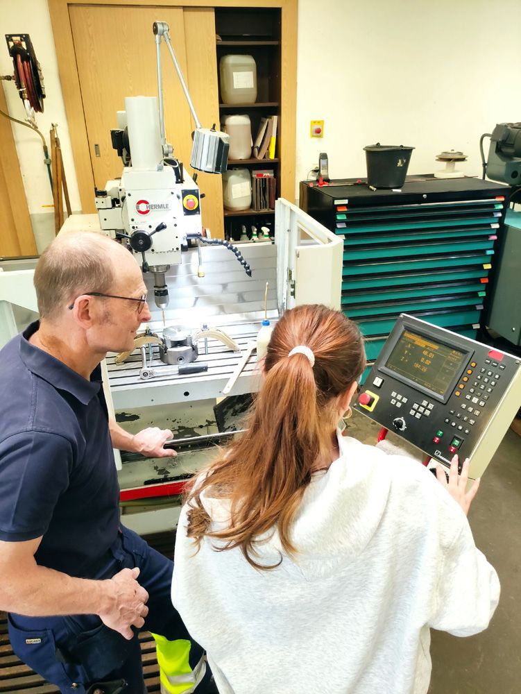Two people reviewing controls beside a machine in a workshop, with tools and equipment in the background.