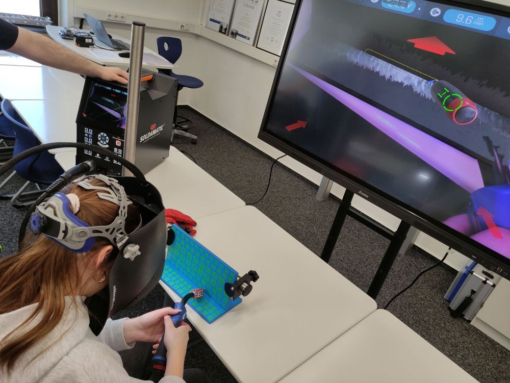 Person in a VR headset using a controller while watching a virtual scene on a large monitor in a lab.