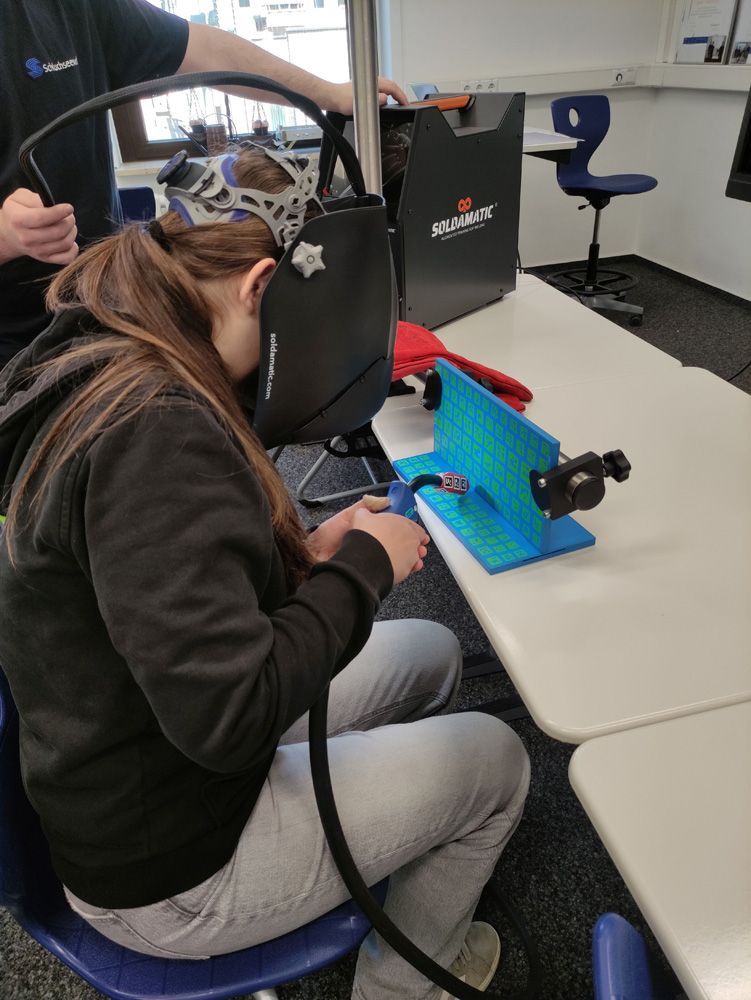 Student wearing a VR headset using a laptop at a classroom desk, with a blue robotic device nearby