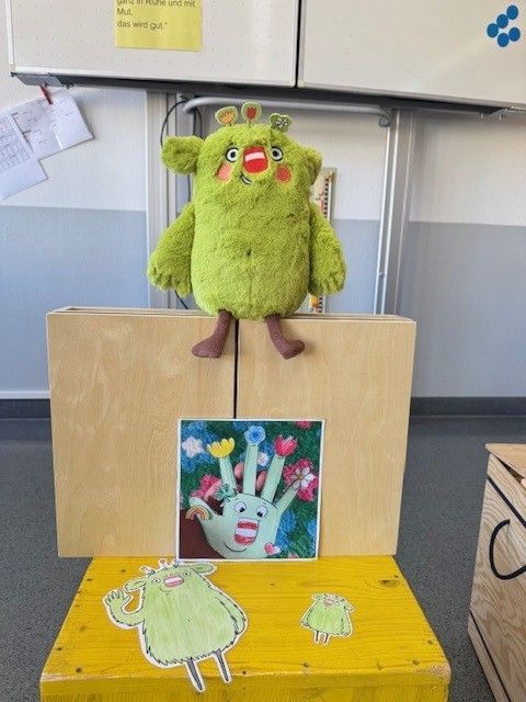 Grüne Plüschmonsterpuppe auf einer Holzbühne mit einem handförmigen Blumenbild.