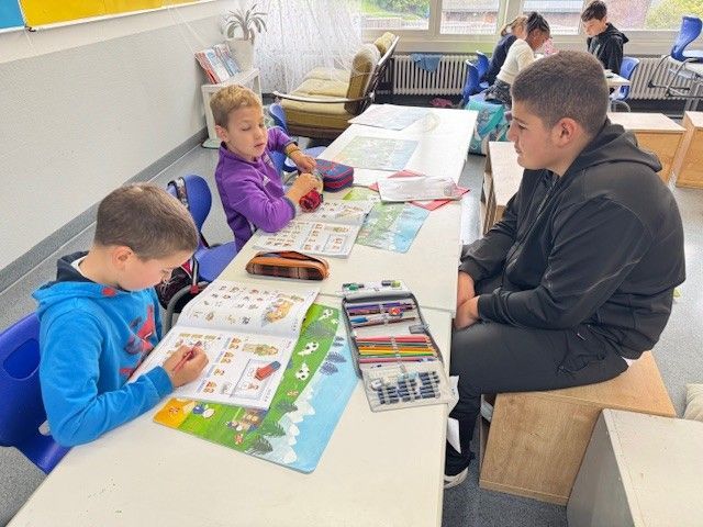 Drei Kinder sitzen an einem Tisch und arbeiten an ihren Aufsätzen. Eines schreibt, ein anderes schaut auf das Papier und das letzte schaut zu ihnen.