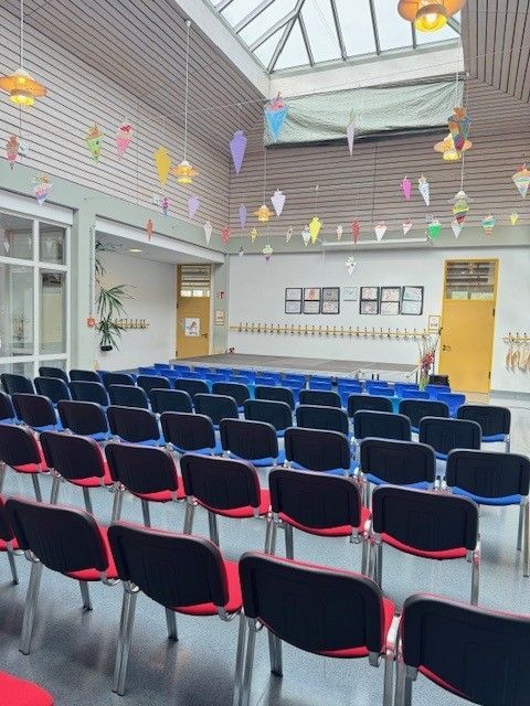 Stuhlreihen in einem Auditorium mit Bühne, von der Decke hängenden Dekorationen und Oberlichtern.