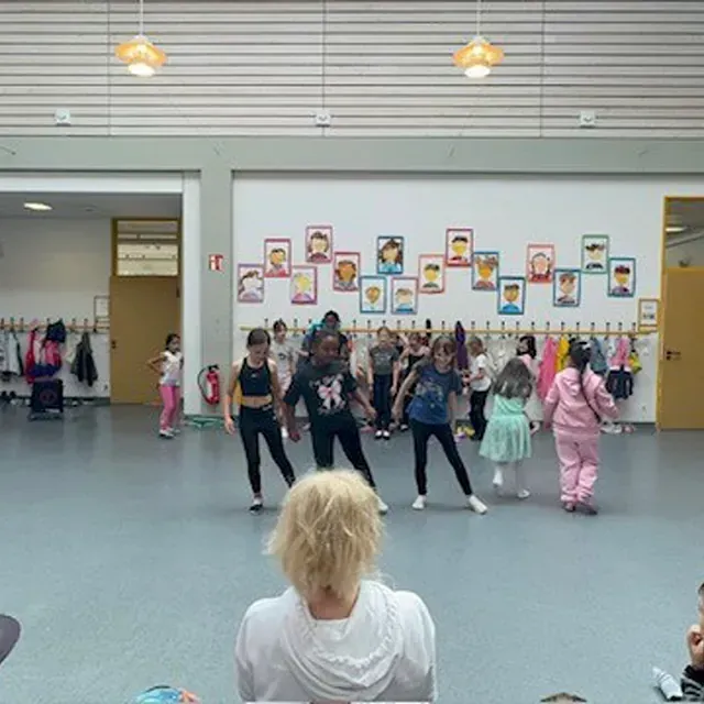 Kinder tanzen in einem Schulflur, die Lehrerin schaut zu. Wand mit Porträts, Kleidung an Haken.