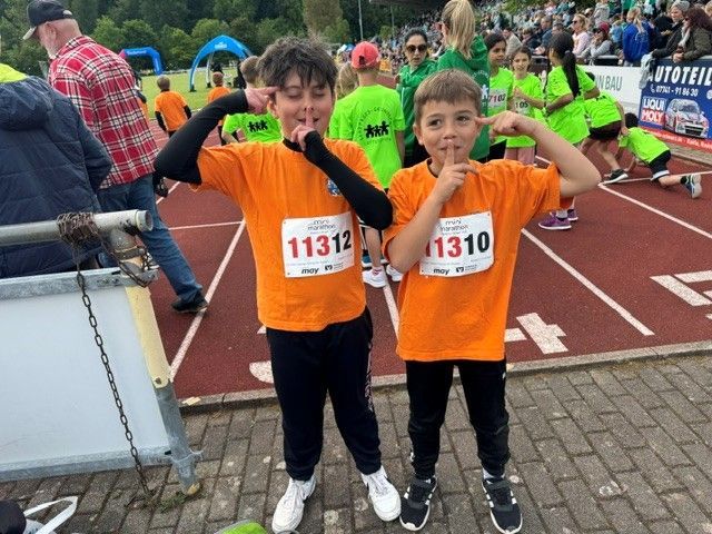 Two children in orange shirts at a race finish line, posing with arms raised and race bibs showing.