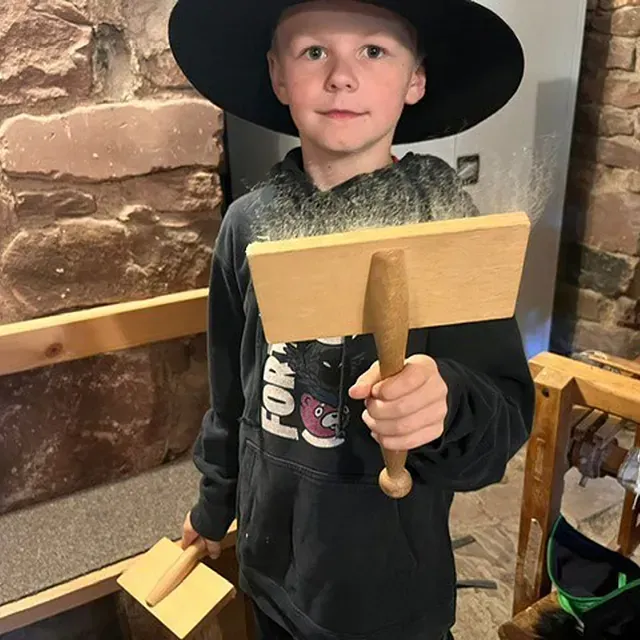 Junge mit Hexenhut, der Holzwerkzeuge hält, steht drinnen in der Nähe einer Steinmauer.