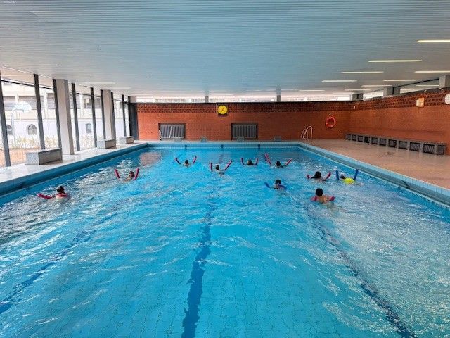 Menschen schwimmen in einem blauen Hallenbad und heben die Arme. Weiße Decke, Backsteinwand, große Fenster.