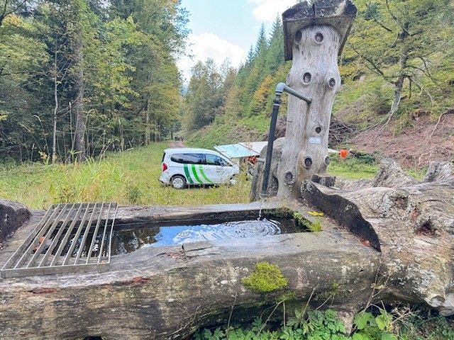 Ein rustikaler, aus Holz gefertigter Brunnen, ein Fahrzeug ist in der Nähe auf einer Rasenfläche geparkt, im Hintergrund sind Bäume zu sehen.
