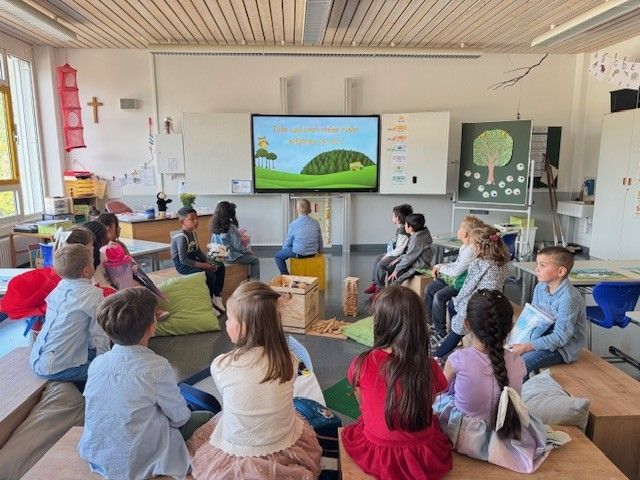 In einem Klassenzimmer sitzen Kinder im Kreis und schauen auf einen Bildschirm. Eine Lehrerin sitzt in der Nähe des Bildschirms.