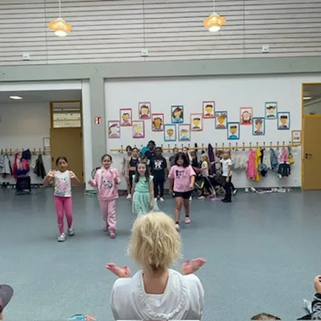 Kinder tanzen in einer Schulturnhalle, beobachtet von einer Lehrerin. Porträts hängen an der Wand, Mäntel reihen sich hinter den Kindern an der Wand.