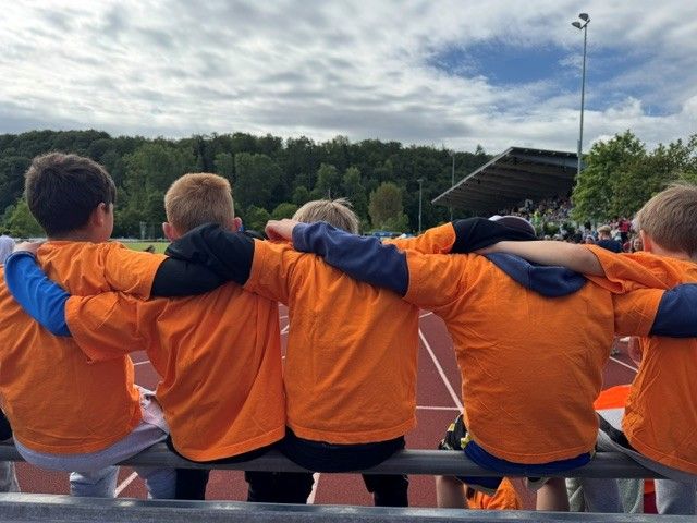 Four people in orange jackets sit arm-in-arm on a bench overlooking a sports field