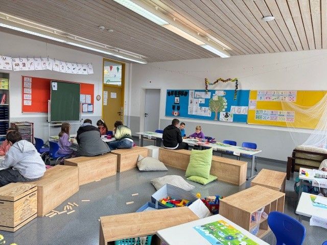 Klassenzimmer mit Kindern und Erwachsenen, die an Tischen und Holzkonstruktionen sitzen und arbeiten.