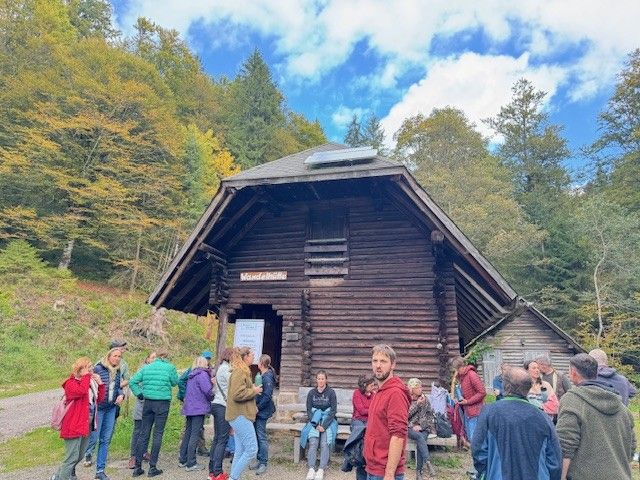 Menschen hatten sich vor einer Holzhütte in einem Wald unter einem teilweise bewölkten Himmel versammelt.