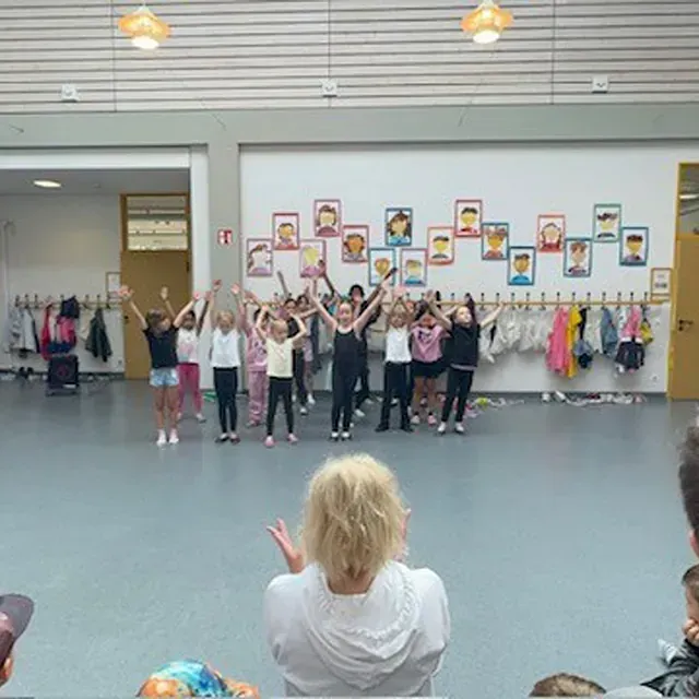 Kinder führen in einem Raum einen Tanz mit erhobenen Armen auf. Eine Frau beobachtet sie aus dem Vordergrund.