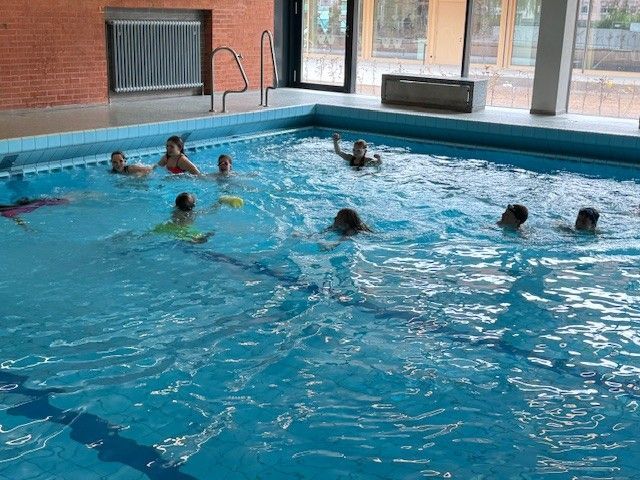 Kinder schwimmen in einem Hallenbad mit blauem Wasser. Einige winken. Backsteinmauer und Fenster sind zu sehen.