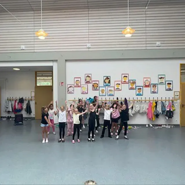 Kinder tanzen mit erhobenen Armen in einem Raum mit Garderobenständern und gerahmten Porträts an der Wand.