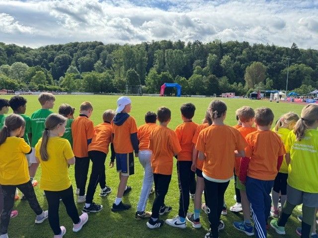 Children in orange and yellow shirts stand on a grassy field near a soccer goal and trees