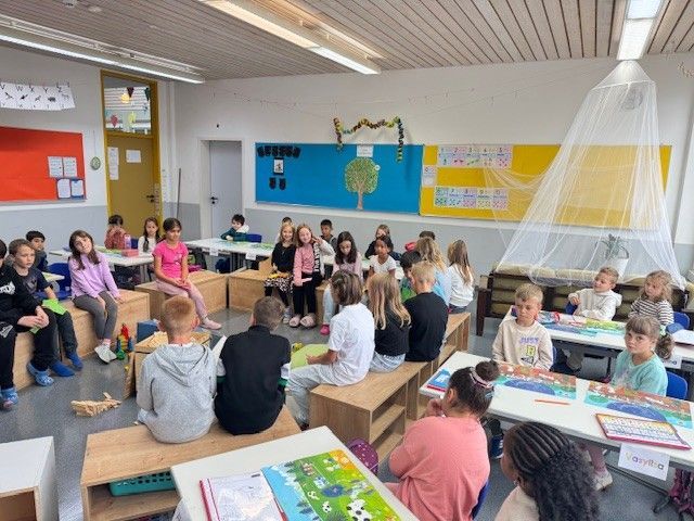 Klassenzimmer mit Kindern, einige sitzen auf Holzklötzen, andere an Tischen. Einige blicken in Richtung eines zentralen Bereichs.