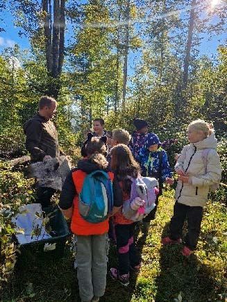 Kinder in orangefarbenen Westen und eine Lehrerin versammeln sich im Freien um einen Baum.