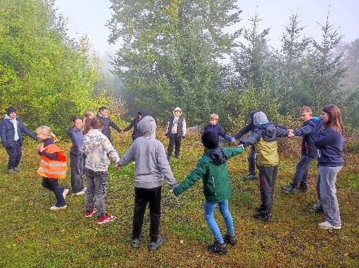 Kinder stehen im Kreis und halten Händchen auf einer Wiese, im Hintergrund Bäume, neblig.