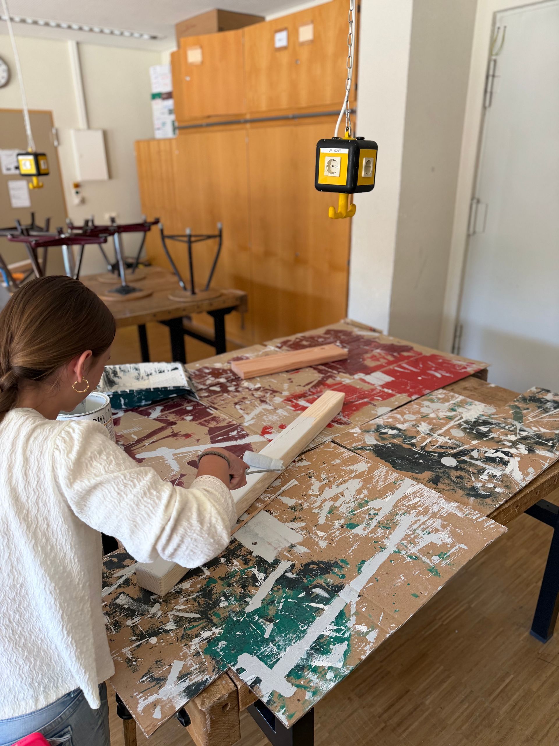 Person painting at a large art table in a studio, with canvases, brushes, and a hanging power tool above.