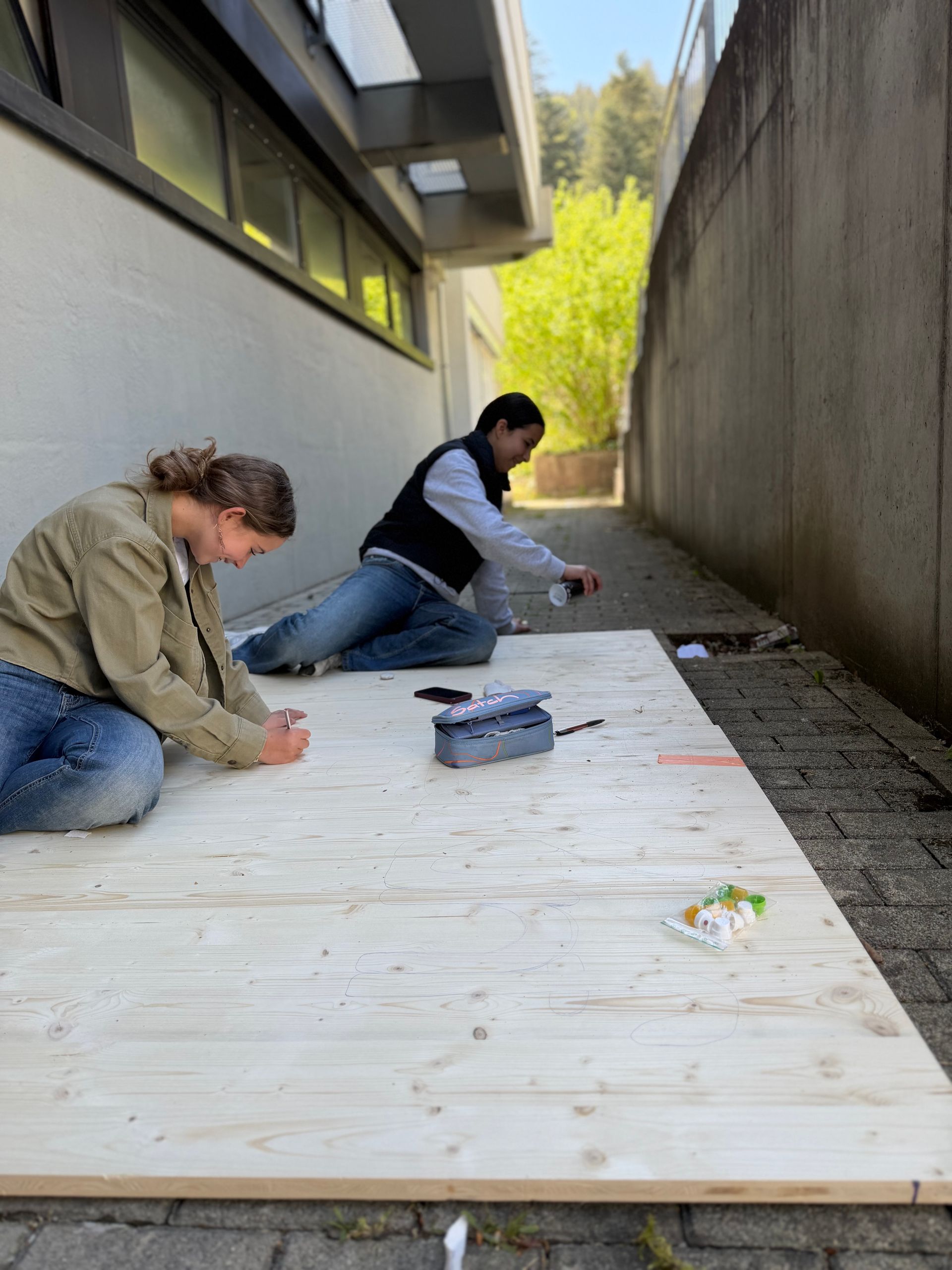 Two people laying a white board walkway beside a building under a sloped concrete wall