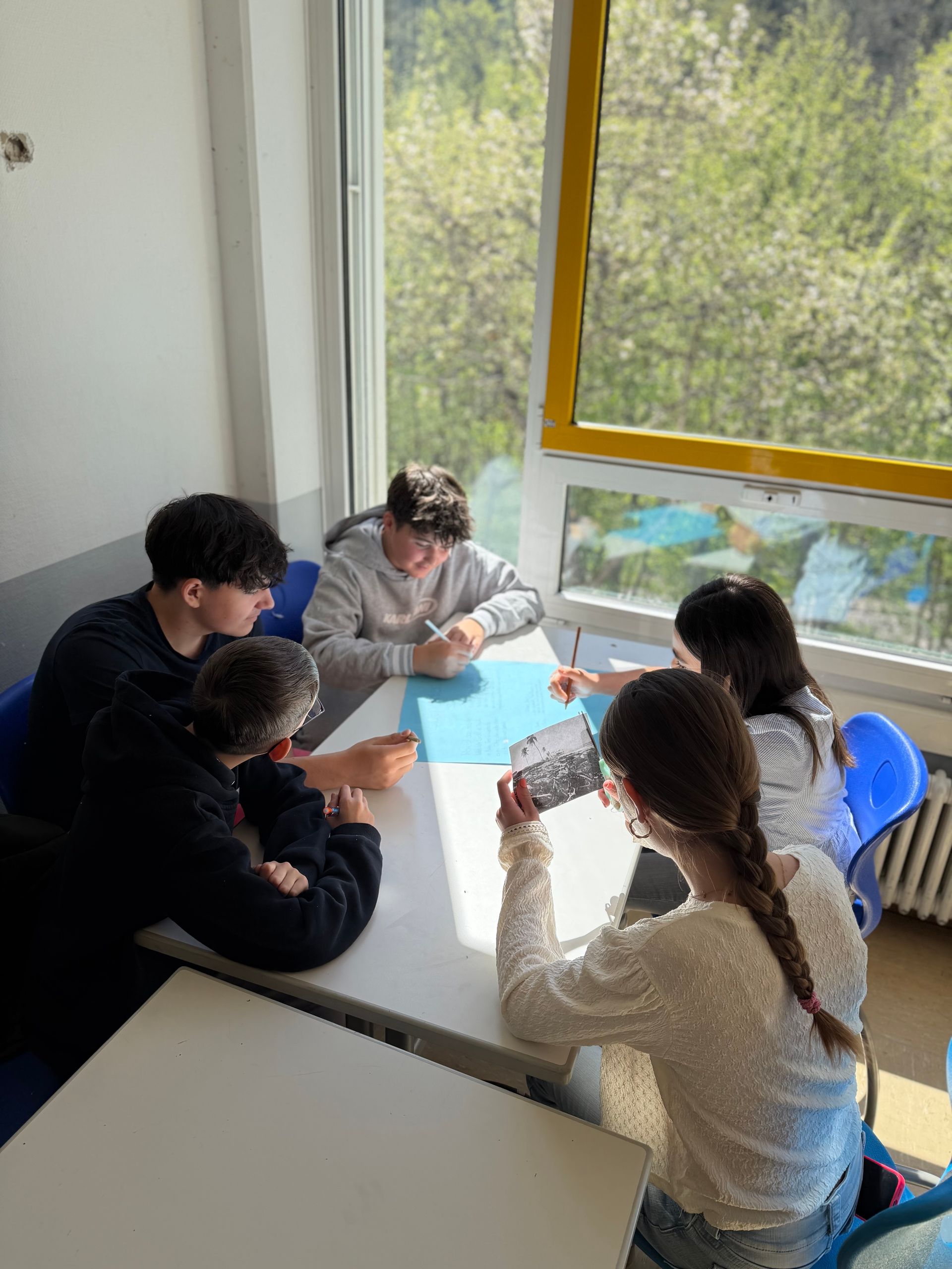 Five people gathered around a table by a bright window, working together on a paper activity.