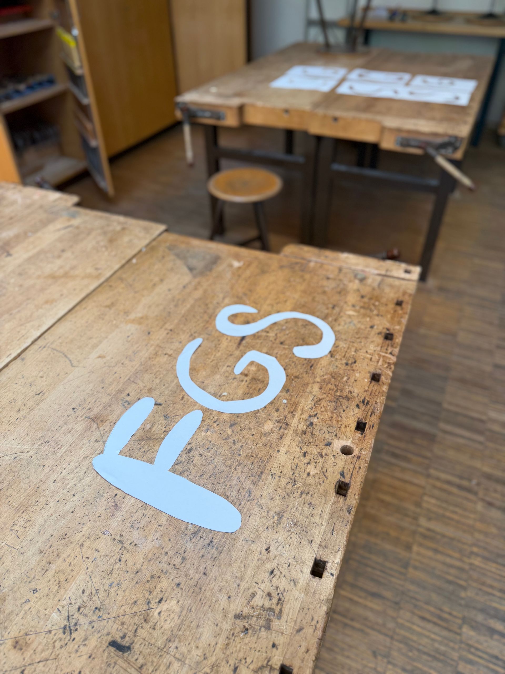 Wooden classroom desks with blue “FCS” lettering, stools, and papers in the background.