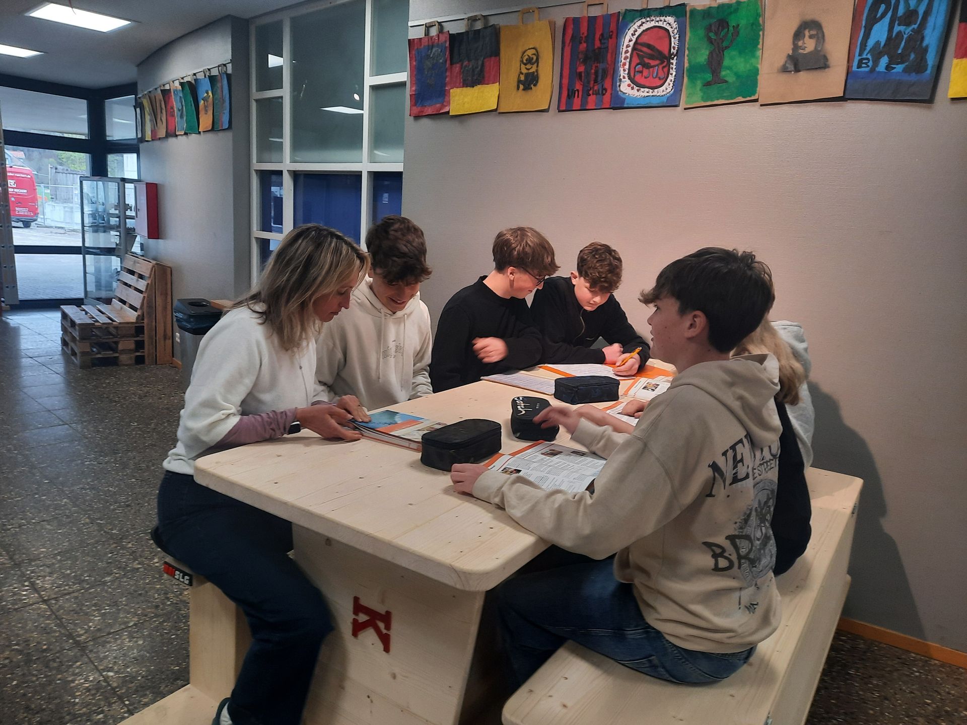 A group of students sits at a wooden table in a school hallway, working on papers together.
