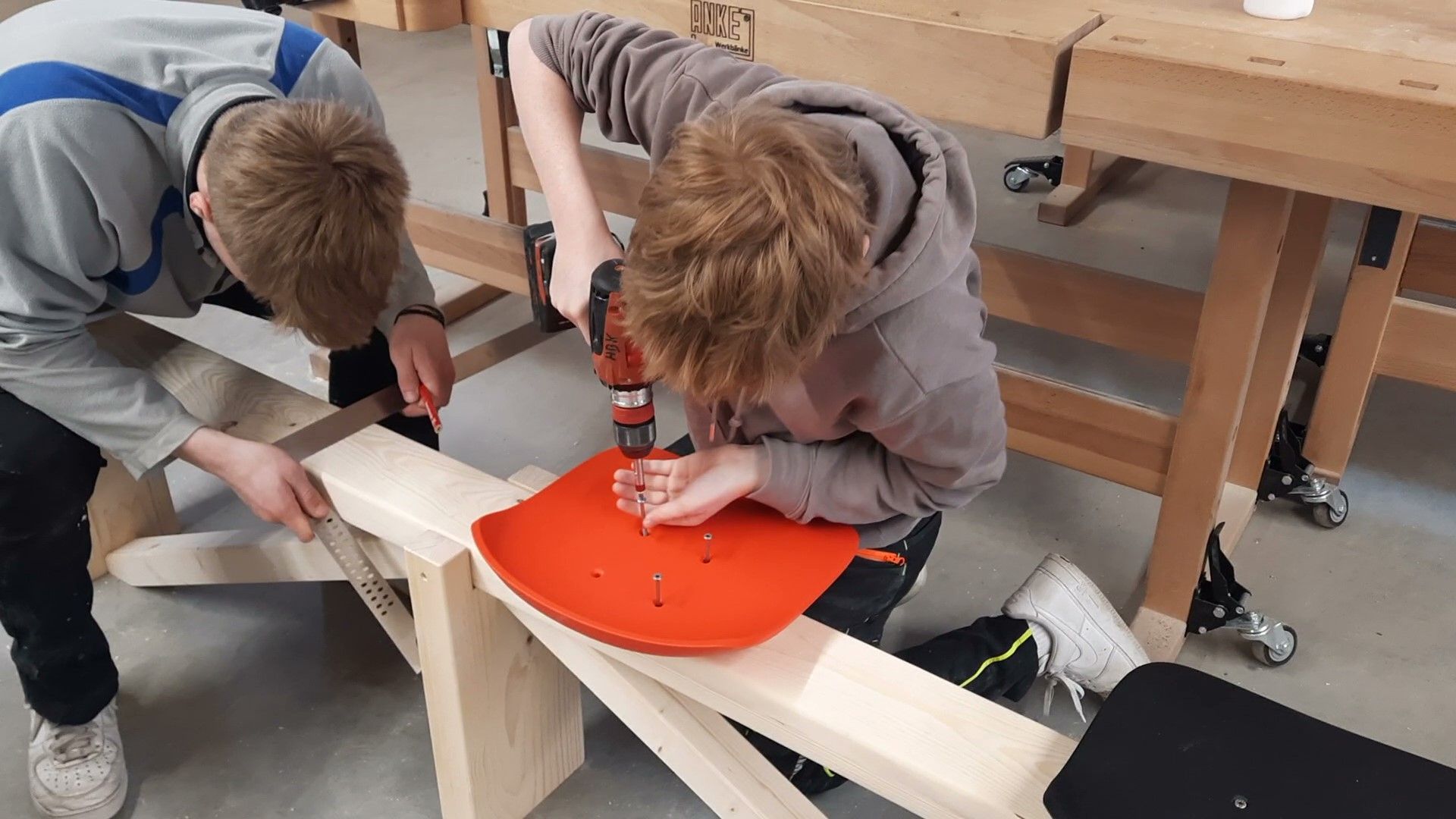 Two people assemble a project on a wooden workbench, one using a power drill on an orange plastic seat.