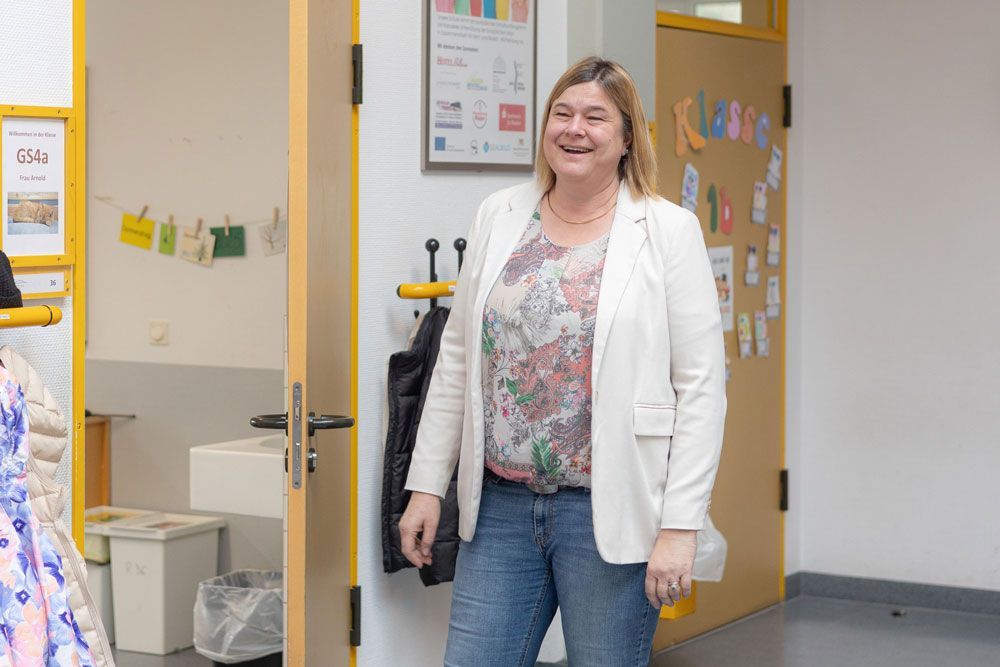 Eine Frau in weißem Blazer und Jeans lächelt in einem hell erleuchteten Flur.