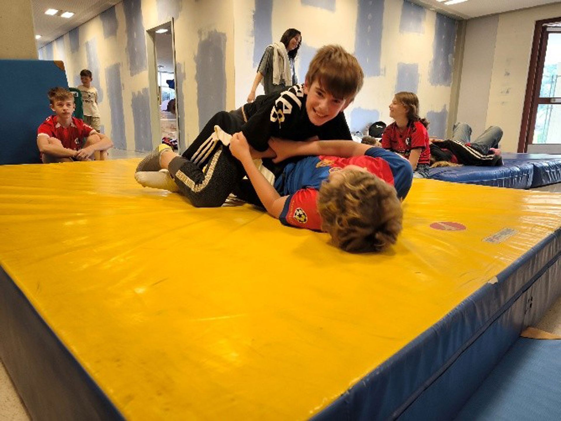Zwei Jungen ringen auf einer gelben Matte in einer Halle, andere schauen zu.