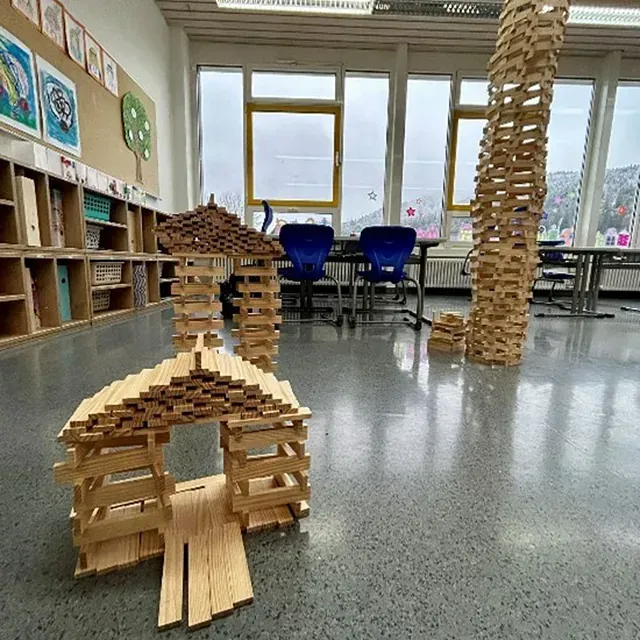 Holzbauwerke in einem Klassenzimmer: ein Haus, ein Turm und ein Torbogen. Im Hintergrund sind ein Fenster und Regale zu sehen.