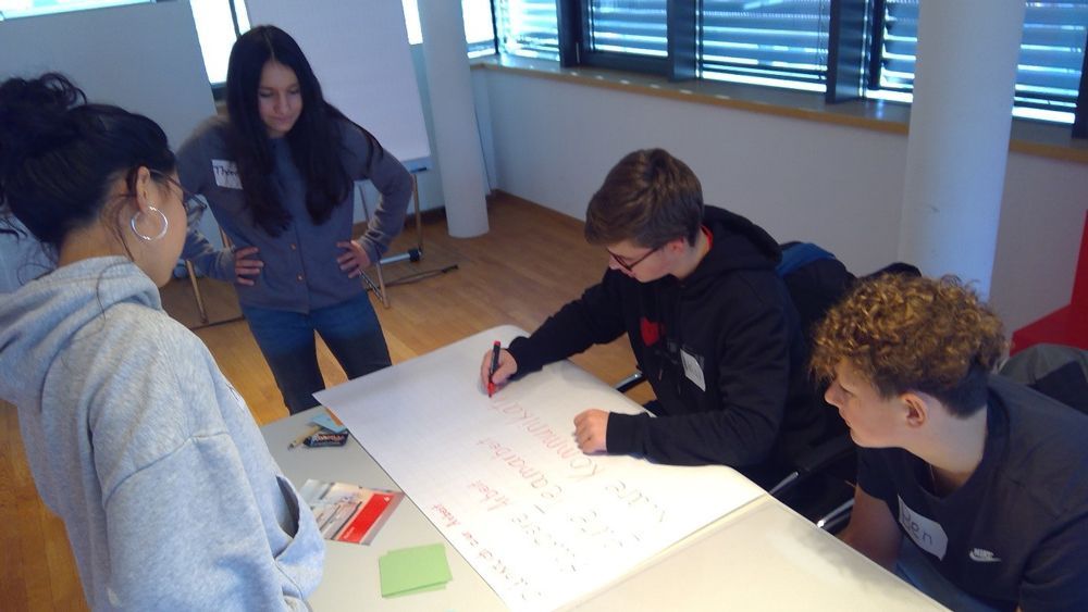 Vier Personen arbeiten in einem hellen Raum an einem Tisch zusammen und schreiben auf einem großen Blatt Papier.