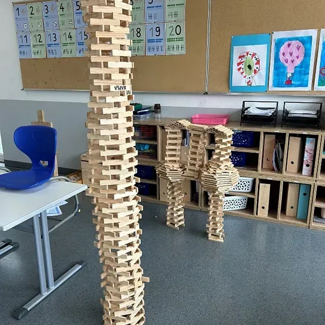 In einem Klassenzimmer mit Zahlentafeln und Kunstwerken befinden sich ein hoher Turm und zwei aus Holzklötzen gebaute Konstruktionen.