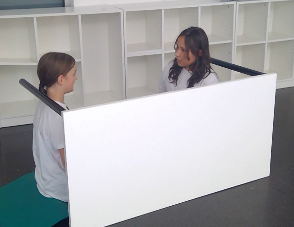 Zwei Personen sitzen sich durch eine weiße Trennwand gegenüber, möglicherweise im Rahmen einer Therapiesitzung, in einem Raum mit Regalen.