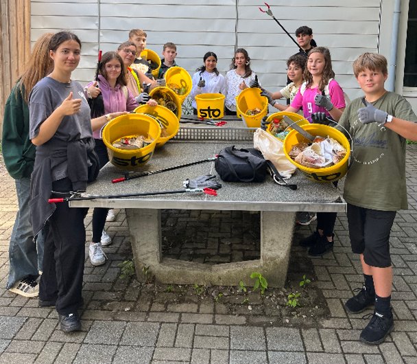 Eine Gruppe Kinder mit Mülleimern und Werkzeugen sitzt um einen Tisch, die Daumen nach oben, im Freien.