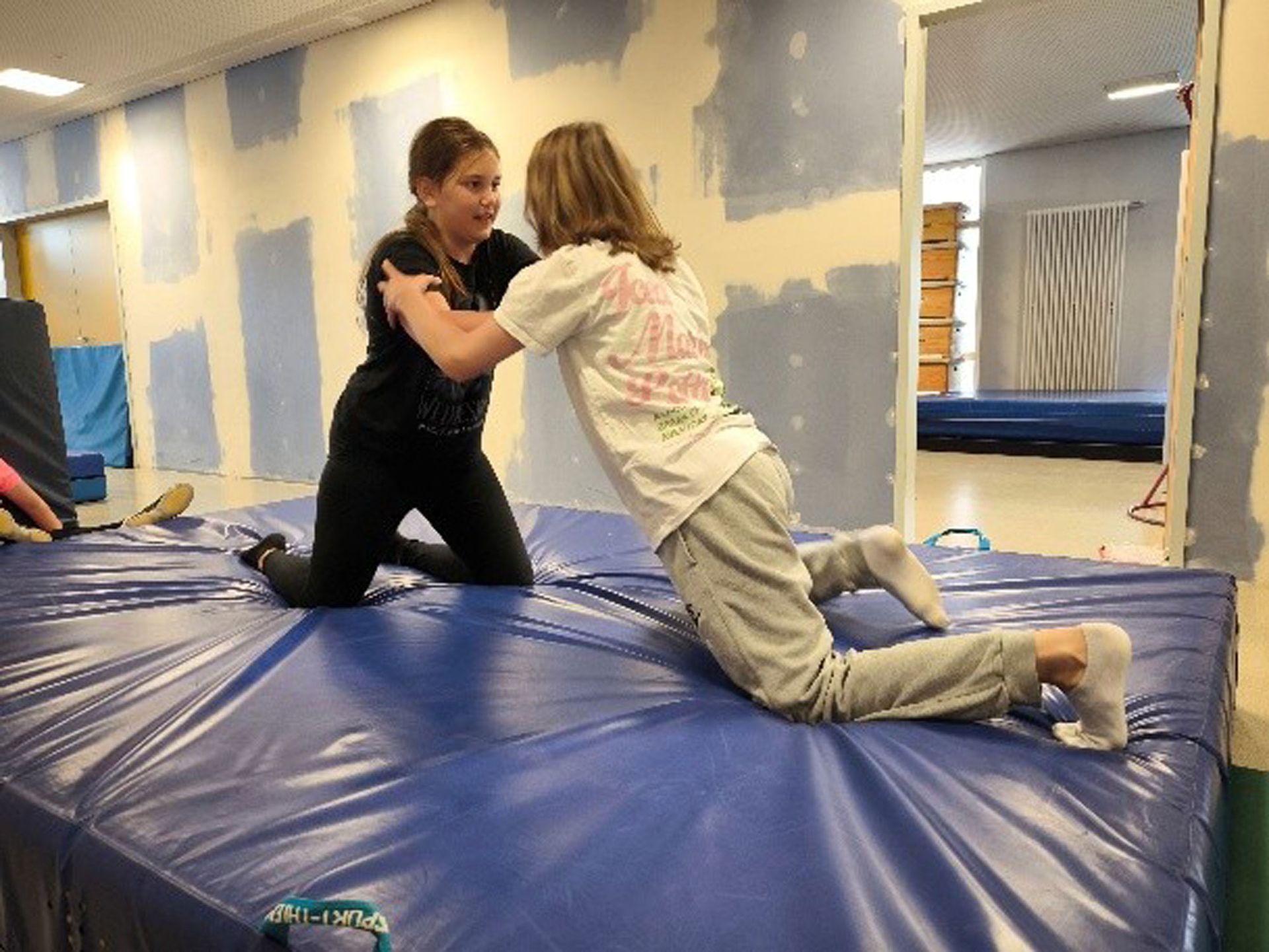 Zwei Mädchen üben Ringen auf einer blauen Matte in einer Turnhalle.