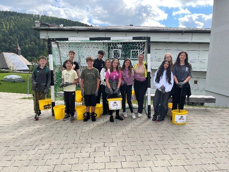Eine Gruppe Jugendlicher mit gelben Eimern posiert im Freien vor einem Tor. Bewölkter Himmel, im Hintergrund ein Gebäude zu sehen.