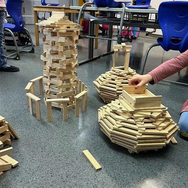 Ein Kind baut mit Holzklötzen auf einem grauen Boden. Ein Turm und zwei bootsähnliche Gebilde sind zu erkennen.