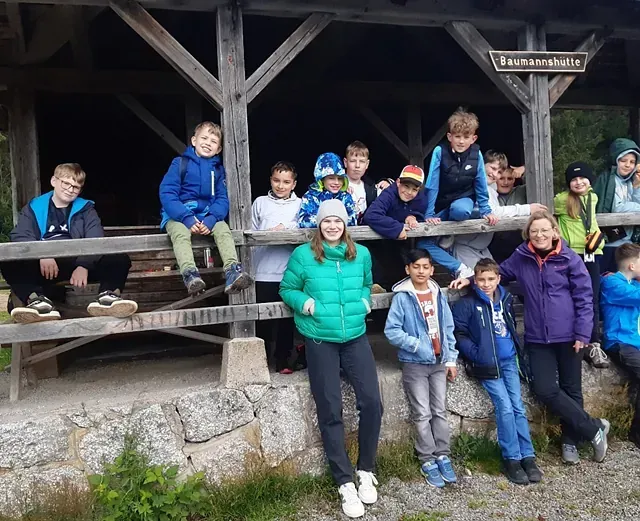 Eine Gruppe von Kindern und zwei Erwachsenen posiert vor einem hölzernen Unterstand; einige sitzen, einige stehen, im Freien.