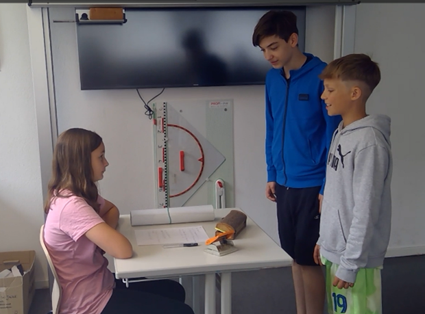 Drei Schüler sitzen an einem Tisch, betrachten eine Zeichnung und unterhalten sich. Einer sitzt, zwei stehen daneben. Ein an der Wand befestigter Bildschirm ist zu sehen.