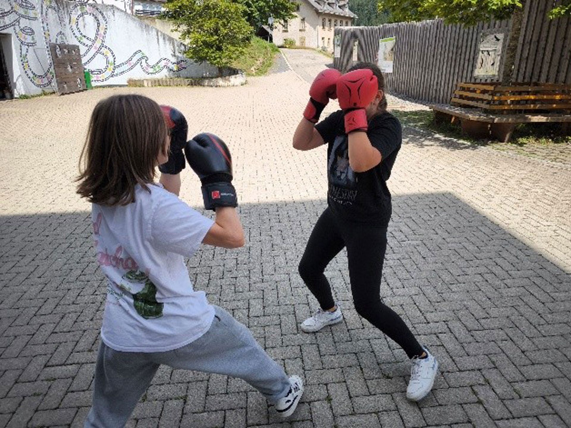 Zwei Personen mit Boxhandschuhen trainieren im Freien. Einer trägt Schwarz, der andere Weiß. Der Platz ist mit Ziegelsteinen gepflastert.
