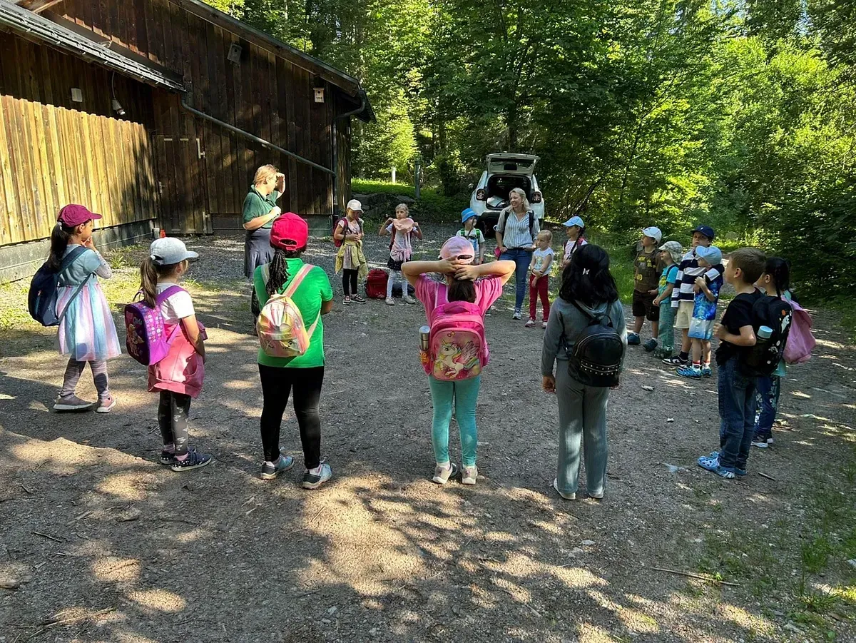 Kinder bei einem Schulausflug stehen im Kreis vor einem Holzgebäude, ihre Rucksäcke sind sichtbar.