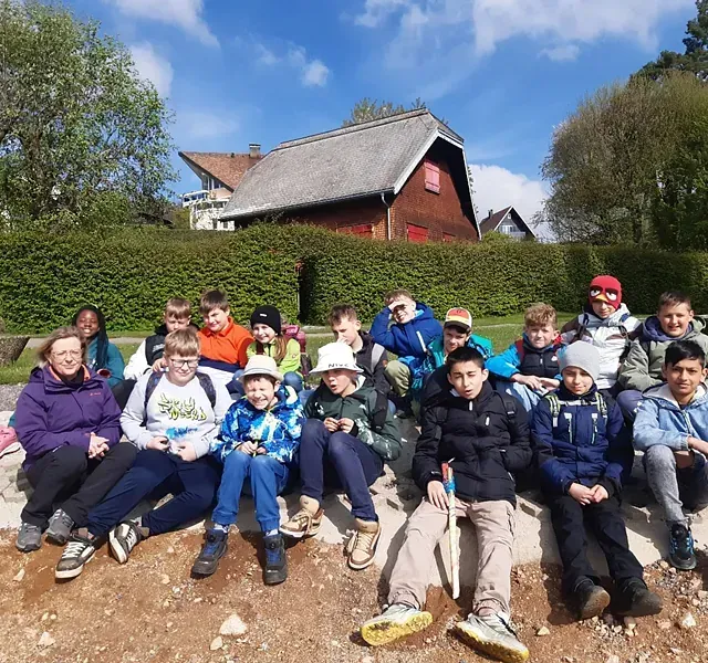 Eine Gruppe lächelnder Studenten sitzt auf einer niedrigen Mauer in der Nähe eines roten Gebäudes unter blauem Himmel.