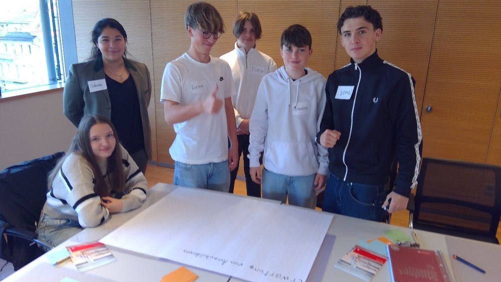 Eine Gruppe von Schülern sitzt um einen Tisch mit einem Poster. Einer zeigt den Daumen nach oben. (Innenraum)