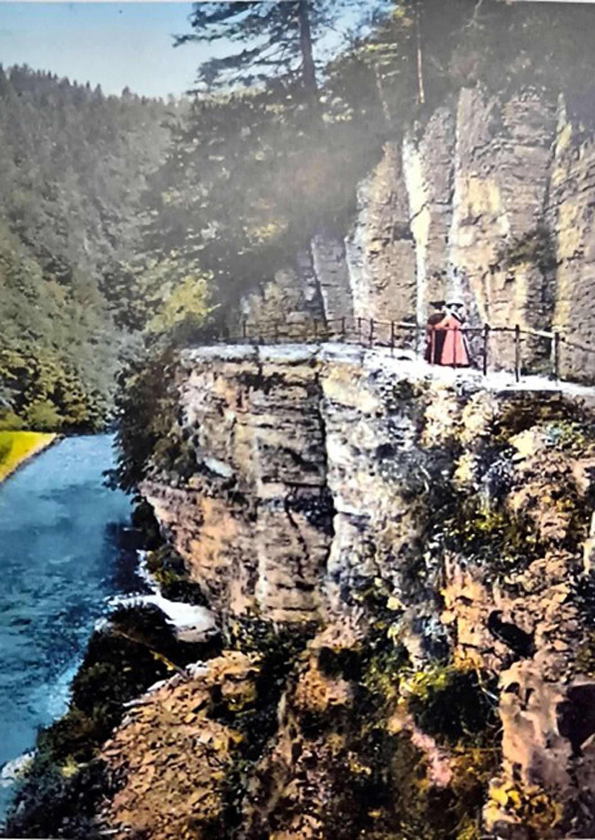 Ein Pfad an einer Klippe entlang eines Flusses. Eine Person in Rot steht in der Nähe des Geländers. Grüne Bäume und blaues Wasser umgeben die Umgebung.