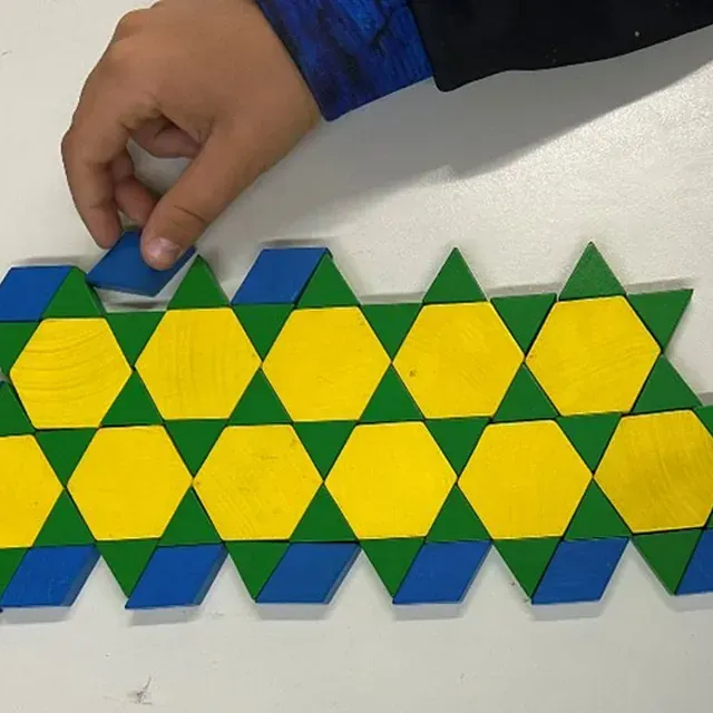 Eine Hand platziert eine blaue Rautenfliese auf einem Mosaikmuster aus gelben Sechsecken, grünen Dreiecken und blauen Rauten.